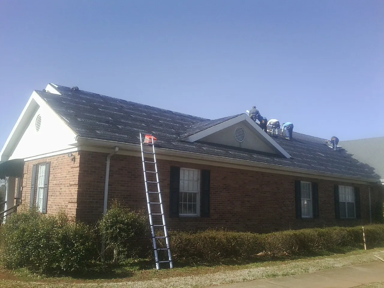 Crew tearing shingles off a brick ranch roof with a ladder set — Emergency Roof Repair work by our crew in Fowlerville