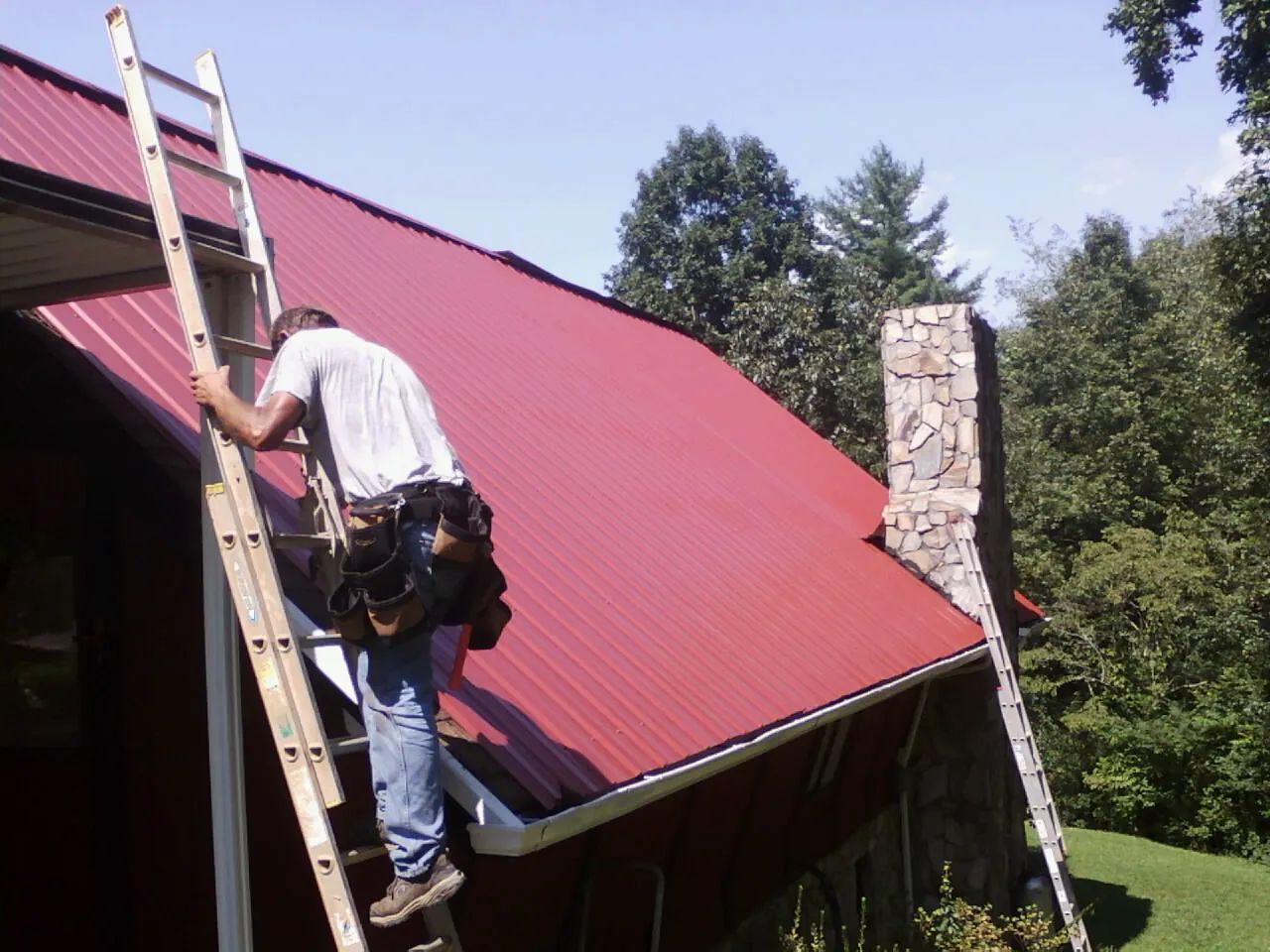 Roofer on a ladder beside a red metal roof and stone chimney — Soffit & Fascia Repair by Roofing Brothers in Fowlerville